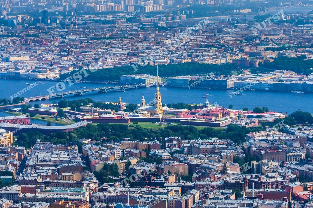На фотографии изображена Петропавловская крепость с её центральным элементом — Петропавловским собором. Снимок сделан с высоты, открывая вид на исторический центр Санкт-Петербурга и извилистую реку Неву. Видны также Троицкий мост и другие знаковые архитектурные сооружения. Яркая зелень и голубые оттенки воды подчёркивают летнюю атмосферу.